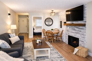 A living room with a fireplace, a TV mounted on the wall, a dining area with chairs, and a sofa with cushions. - Walk to the Strip • Mountain Views • Cozy Retreat (Gatlinburg)