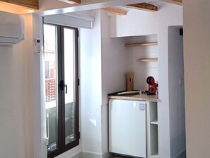 Une cuisine compacte équipée d’un réfrigérateur, d’une fenêtre donnant sur des immeubles et d’une porte menant à un espace extérieur.
