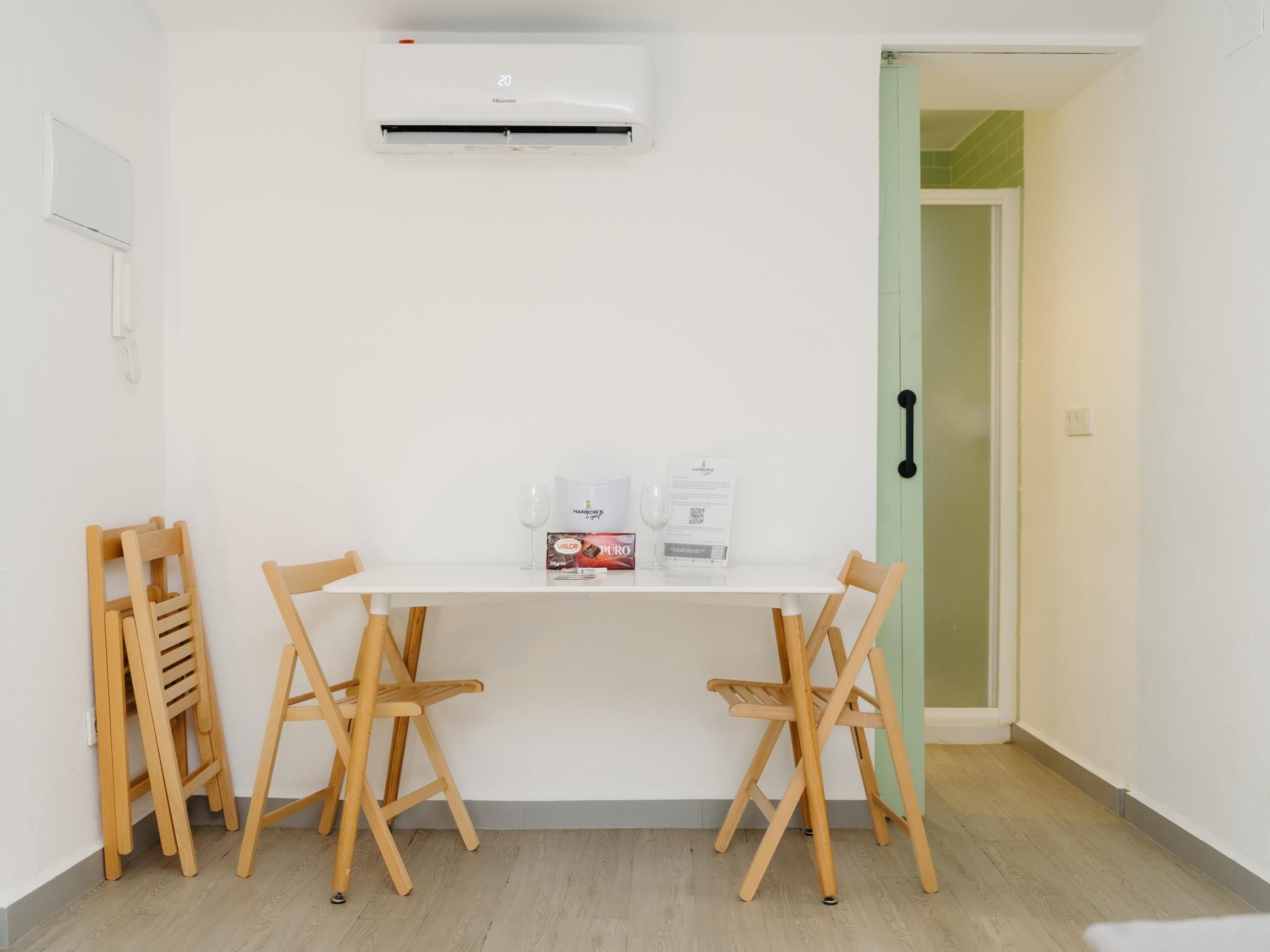 Una habitación pequeña y limpia con una mesa blanca, dos sillas de madera, un equipo de aire acondicionado blanco y una puerta con marco verde.