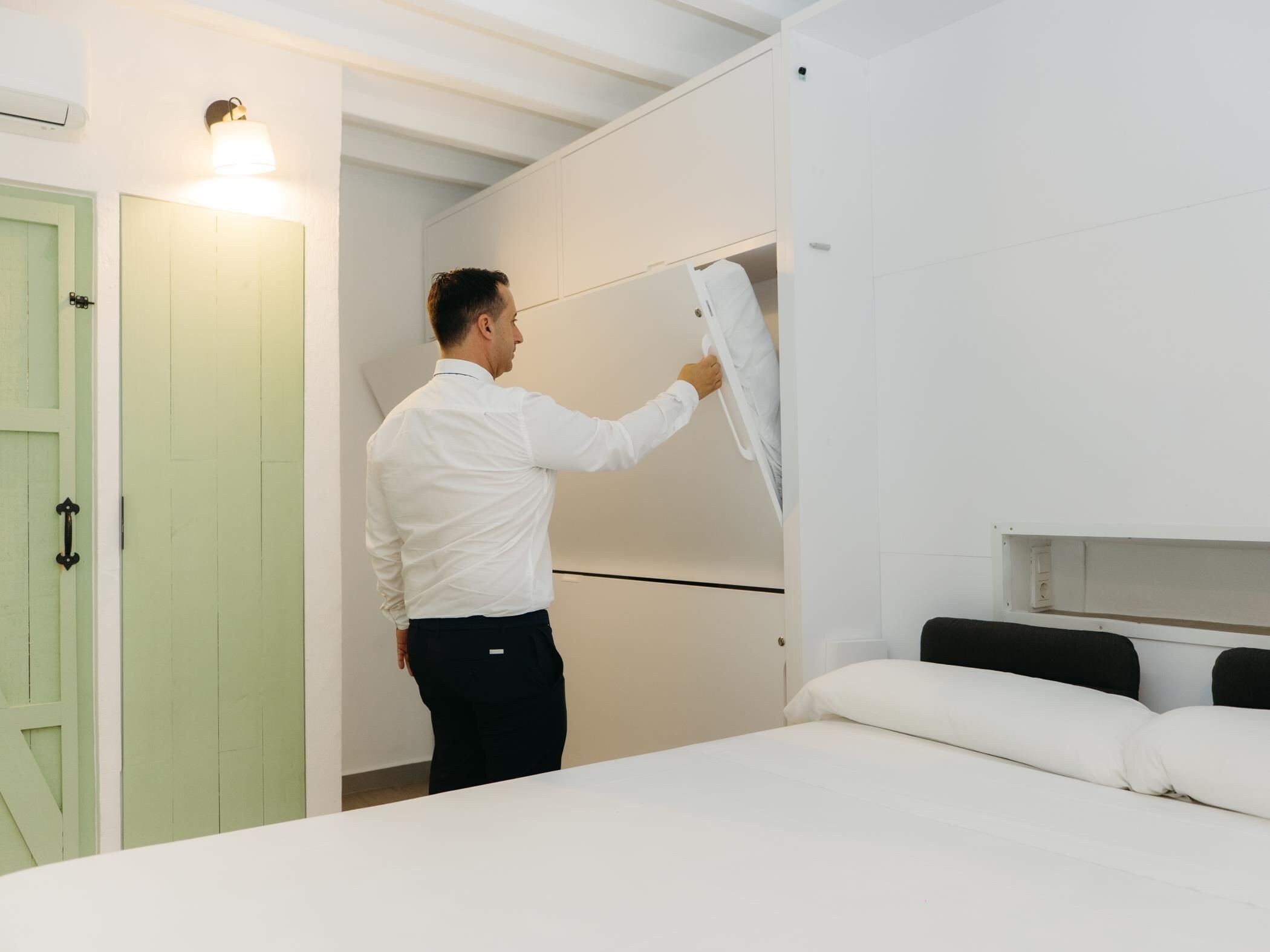 En person installerer en hvid, vægmonteret enhed i et rum med en seng, en grøn dør og et hvidt loft.