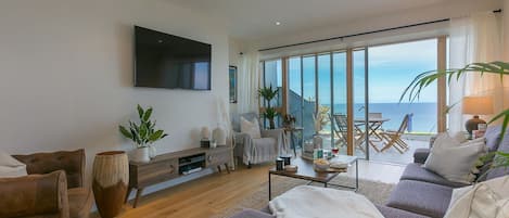 A modern living room with a large TV, comfortable seating, a coffee table, and a view of the sea.