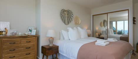 A bedroom with a bed, bedside tables, a dresser, a mirror, and a view of the sea.