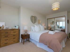 A bedroom with a bed, bedside tables, a dresser, a mirror, and a view of the sea. - Anchors Rest (St Ives)