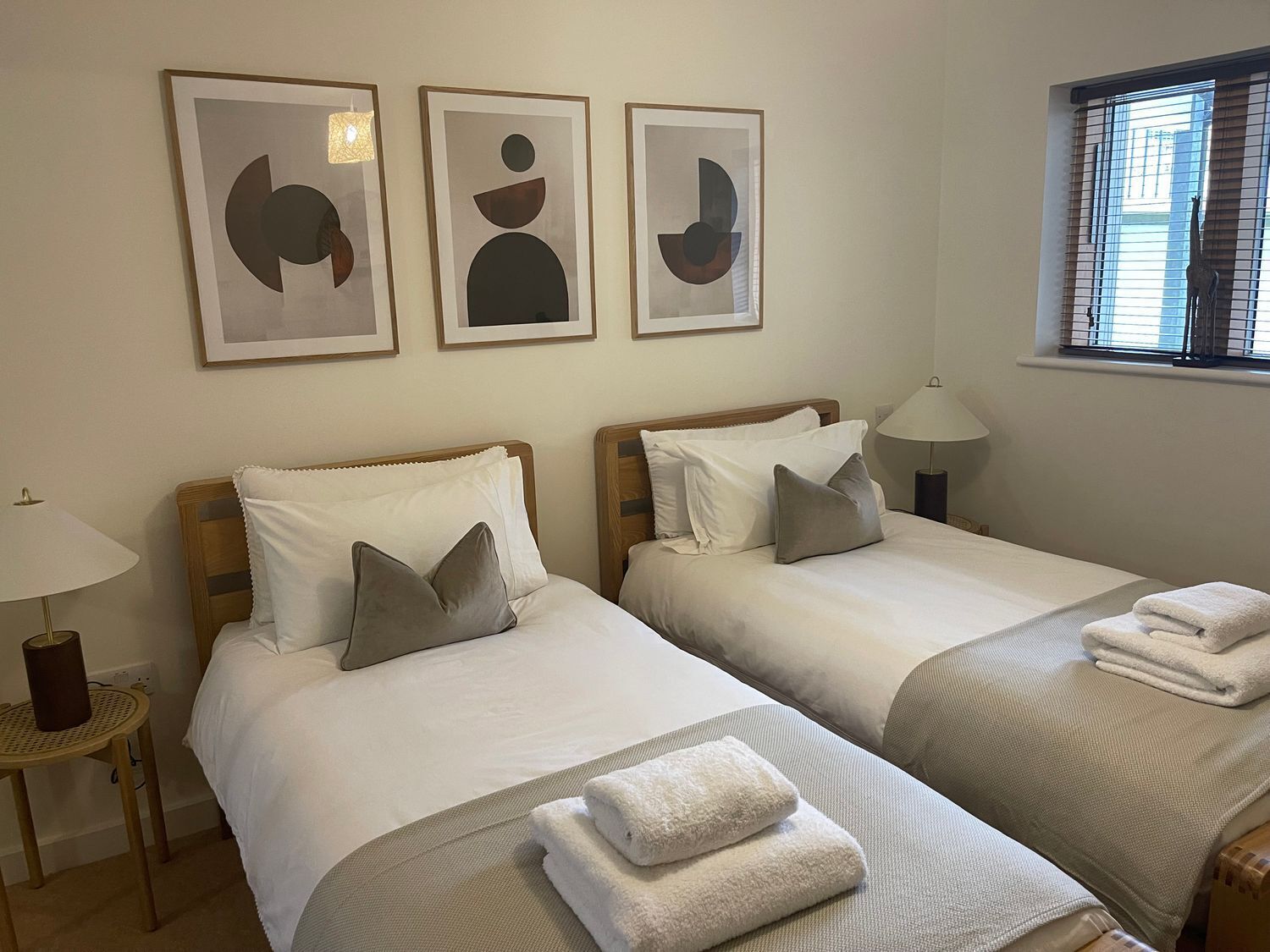 Habitación de hotel con dos camas, una ventana con persianas, una lámpara y una obra de arte abstracta en la pared.