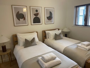 A hotel room with two beds, a window with blinds, a lamp, and abstract artwork on the wall. - Anchors Rest (St Ives)