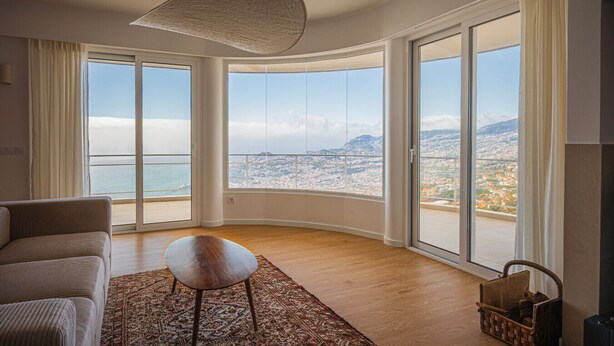 Ein geräumiges Wohnzimmer mit großem Fenster und Panoramablick auf die Stadt, einer Couch, einem kleinen Tisch und einem gemusterten Teppich.