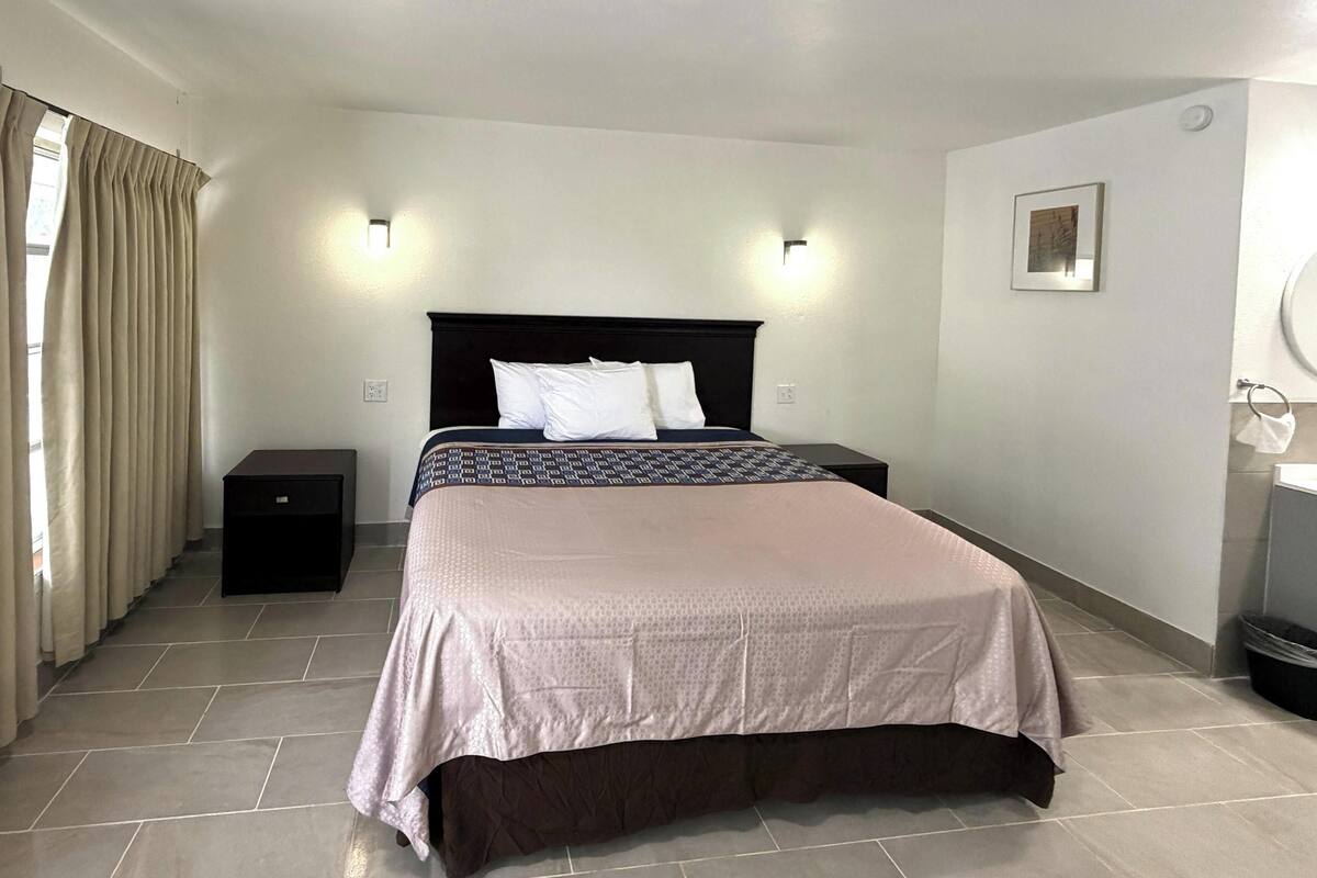 A hotel room with a bed, bedside tables, a wall-mounted light, a framed picture, and a window with curtains.
