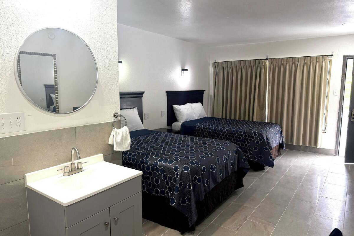 A hotel room with a round mirror, two beds with patterned bedspreads, a white sink, and a window with curtains.