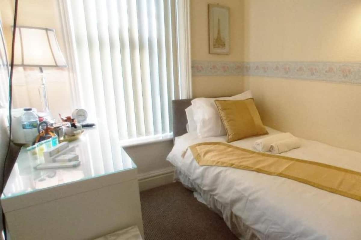 A small hotel room with a single bed, a bedside table with toiletries, a window with blinds, and a wall with a decorative border.