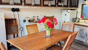 Une table de salle à manger en bois, entourée de chaises, un vase rempli de fleurs et, au fond, un coin cuisine avec des placards et des appareils électroménagers.