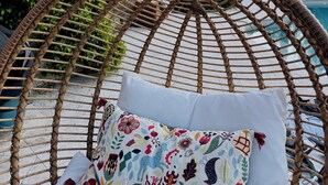 Un fauteuil suspendu en osier avec un coussin à motifs floraux, une piscine et une végétation luxuriante en arrière-plan.