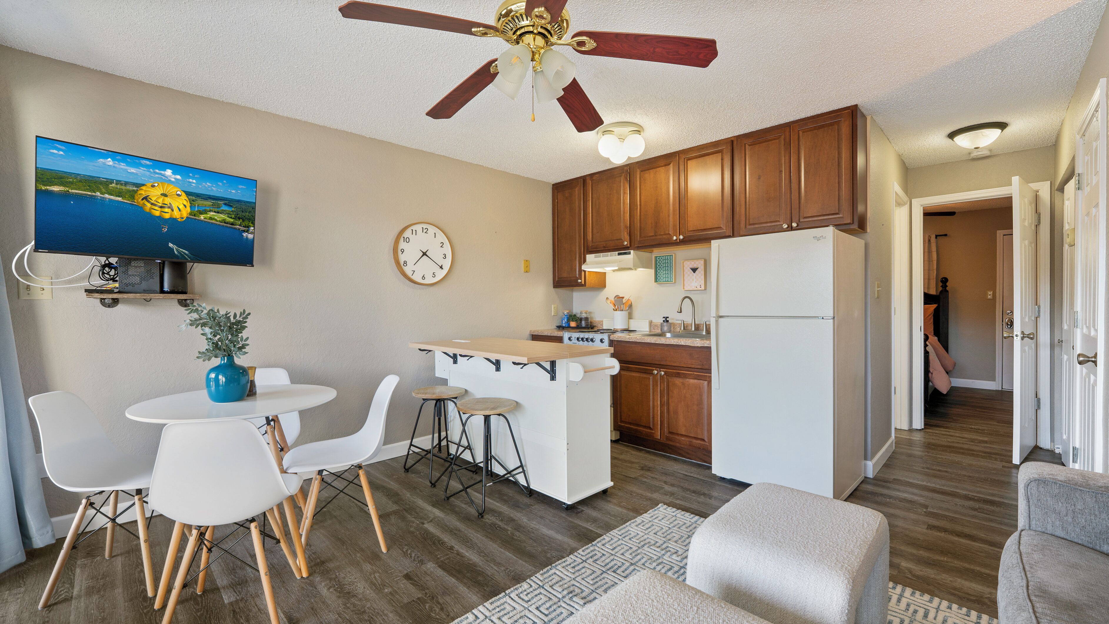 A compact kitchen with wooden cabinets, a built-in stove, and a refrigerator. There is a dining area with white chairs and a small table. A ceiling fan and a clock are visible.