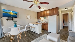 A compact kitchen with wooden cabinets, a built-in stove, and a refrigerator. There is a dining area with white chairs and a small table. A ceiling fan and a clock are visible. - Cozy Lake Escape - King Bed • Private Deck (Osage Beach)
