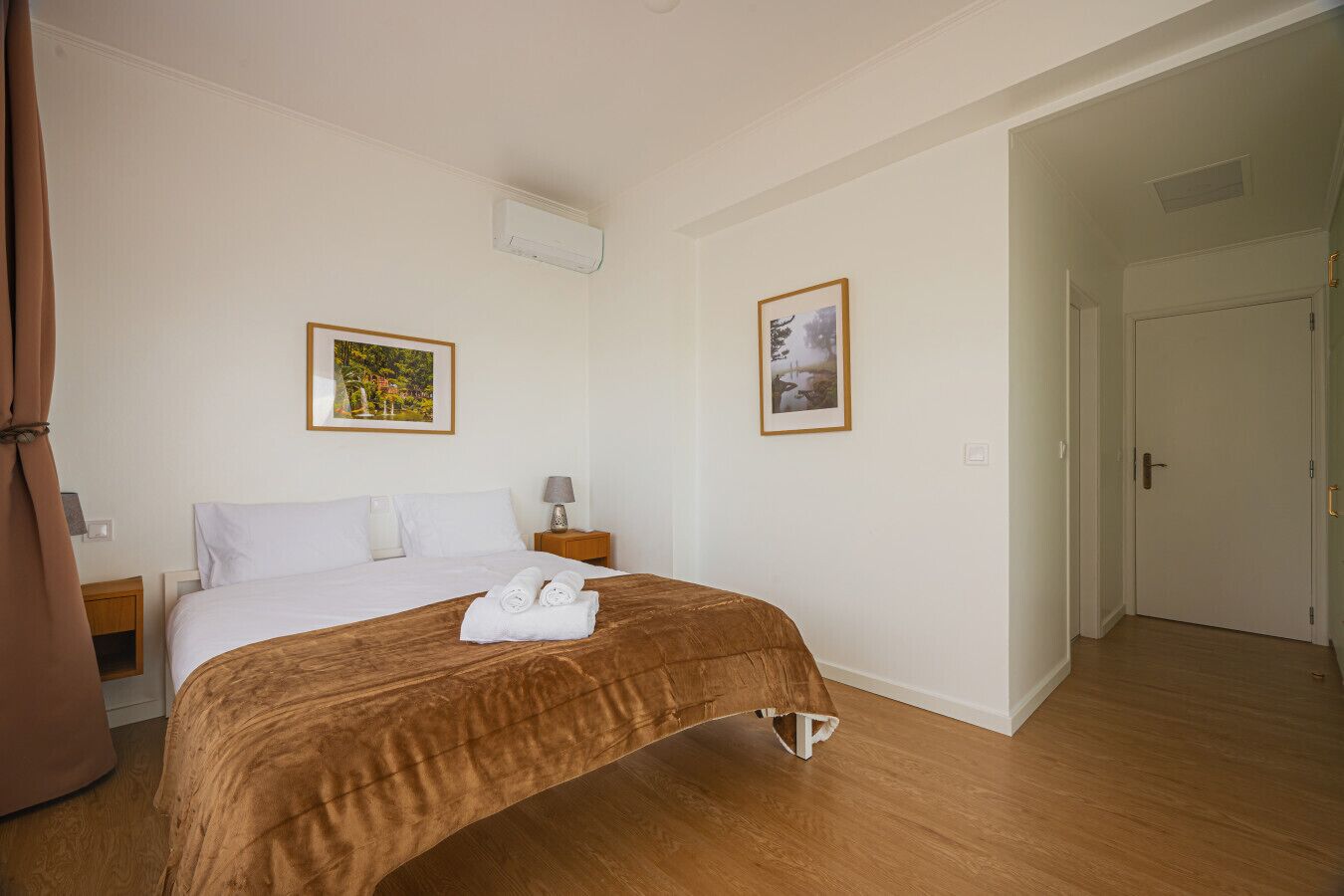 A hotel room with a bed, two framed pictures on the walls, a bedside table with a lamp, and a door leading to another room.