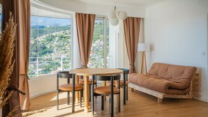 A room with a wooden floor, a round table with chairs, a sofa, and a view of a town through large windows. - 2 Bedroom | 180° View + Balcony | Bathtub (Funchal)