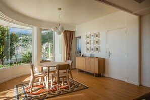 A room with a dining table, chairs, a rug, a cabinet, and a view of a town through large windows. - Panoramic Ocean View | Pool & Garden | 2 Bedrooms (Funchal)