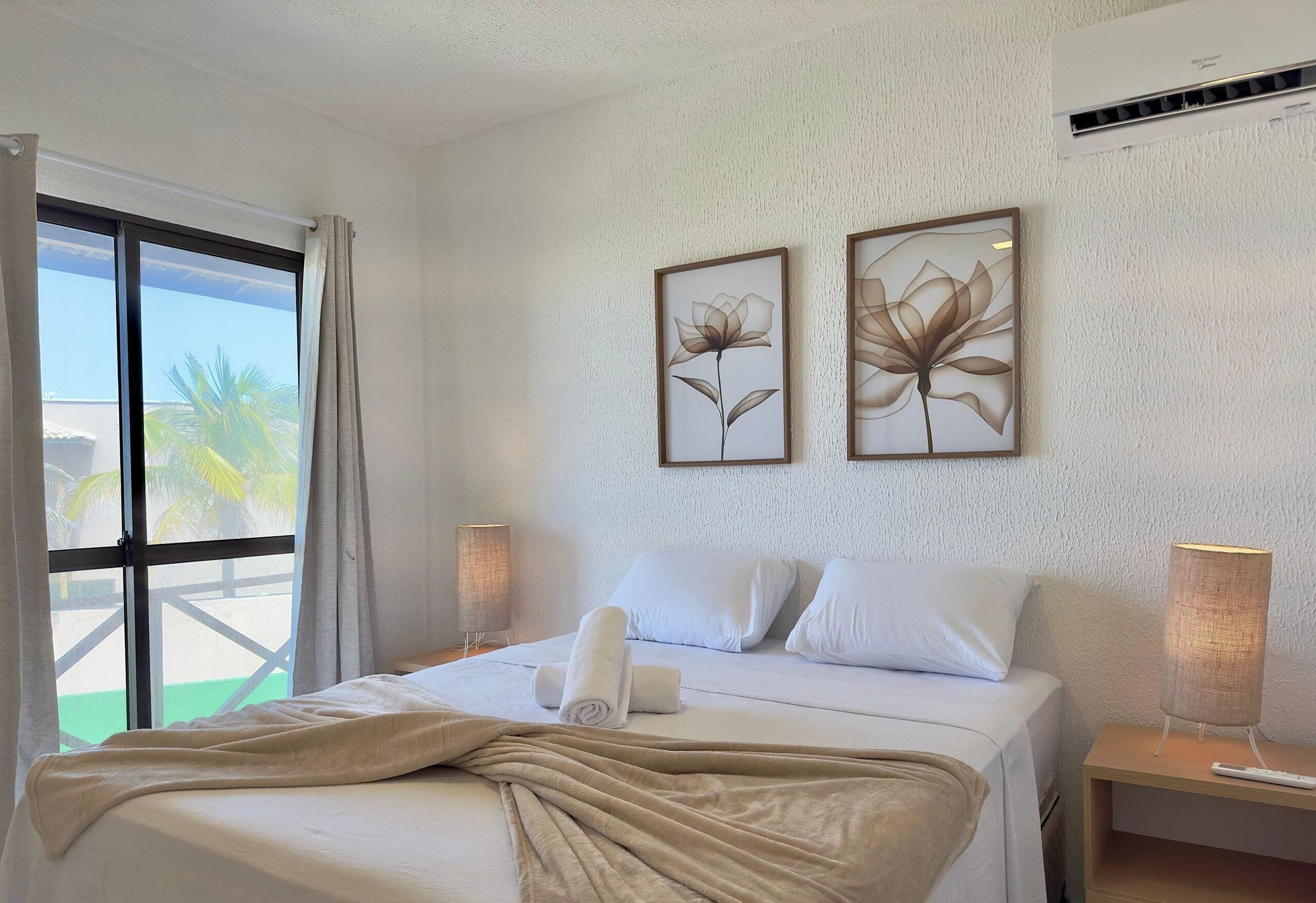 Une chambre d’hôtel avec un lit, deux tableaux représentant des fleurs accrochés au mur, une fenêtre avec des rideaux, une lampe de chevet et un appareil de climatisation.