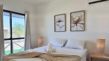 A hotel room with a bed, two wall-mounted flower paintings, a window with curtains, a bedside lamp, and an air conditioning unit.