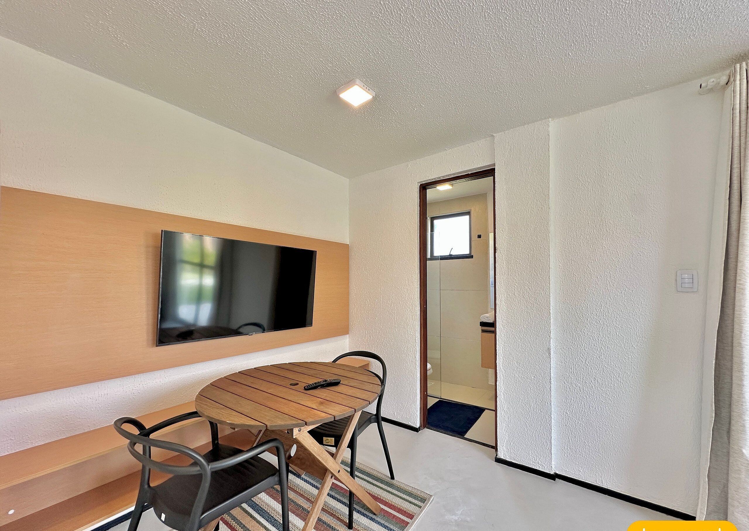 Una habitación moderna con un televisor de pantalla plana montado en un panel de madera, una mesa redonda de madera con sillas y una puerta que da al baño.
