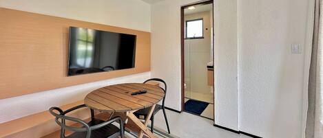 Una habitación moderna con un televisor de pantalla plana montado en un panel de madera, una mesa redonda de madera con sillas y una puerta que da al baño.