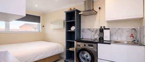 Habitación de hotel pequeña con cama, lavadora, lavamanos y una kitchenette.