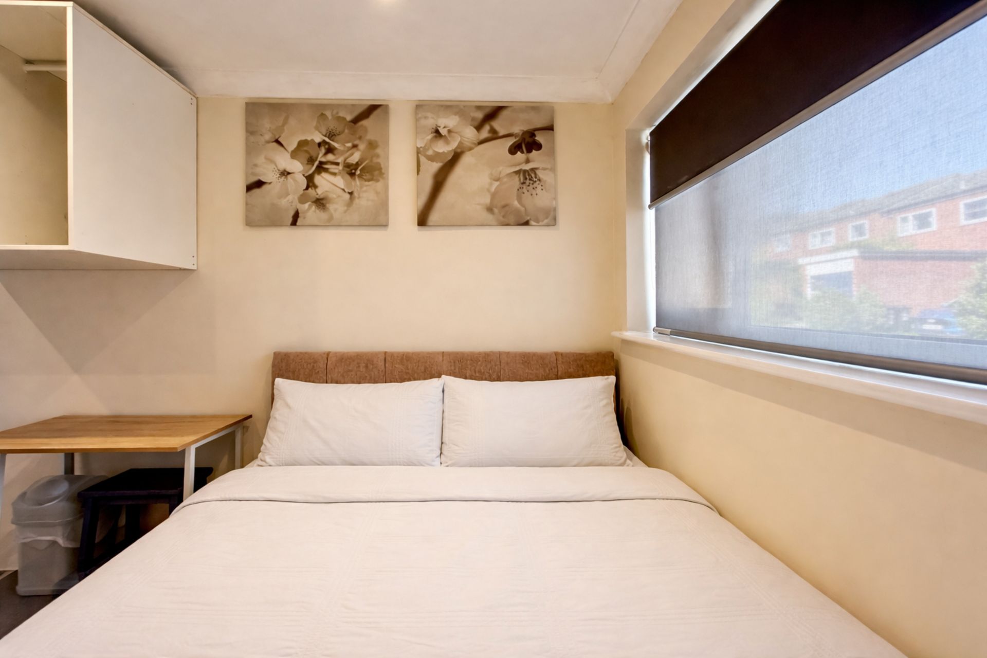 A single-bed room with a wooden headboard, a small table, a trash bin, and two wall-mounted flower paintings.