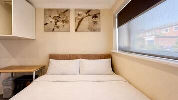 A single-bed room with a wooden headboard, a small table, a trash bin, and two wall-mounted flower paintings.