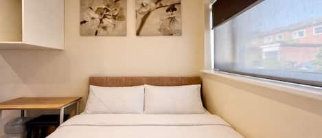 Habitación con una cama individual, cabecera de madera, una mesita, un cesto de basura y dos cuadros de flores colgados en la pared.
