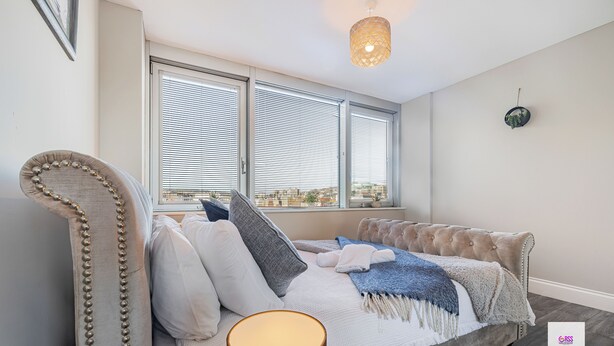 A bedroom with a tufted headboard, a bed with a white and blue blanket, two windows with blinds, a hanging light fixture, and a small wall decoration. - Brighton Central Seaview Retreat, Steps to the Beach (Brighton and Hove)