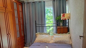 A bedroom with a bed, curtains, a window with a view of greenery, a bedside table with a lamp, and a wardrobe.