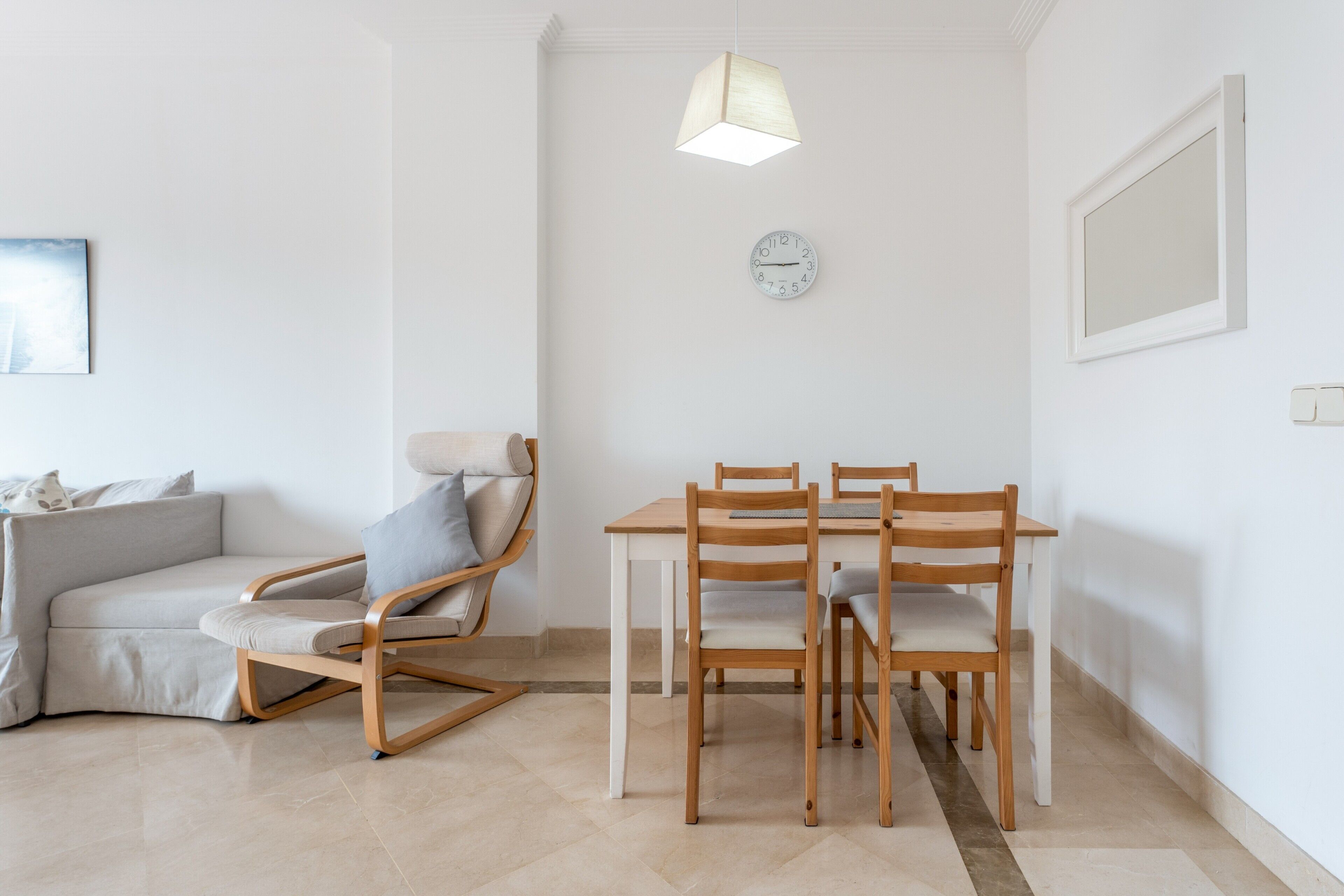 A modern living room with a sofa, a wooden dining table with chairs, a hanging light fixture, and a wall clock.