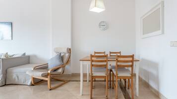 A modern living room with a sofa, a wooden dining table with chairs, a hanging light fixture, and a wall clock.
