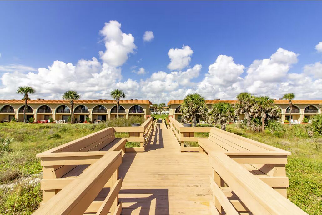 Une promenade en bois mène à une rangée de bâtiments dotés d’arcs en plein cintre et bordée de palmiers.