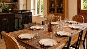 Mesa de jantar posta com pratos, copos e talheres, cadeiras de madeira, um vaso com flores secas e uma área de cozinha com geladeira e armários.