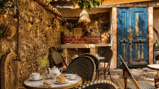 Um café rústico ao ar livre, com paredes de pedra, móveis de vime e uma porta azul.