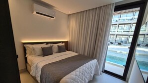 A modern hotel bedroom with a large bed, a ceiling air conditioner, and a view of a pool and building through a glass door. - Porto 2 Life Resort (Ipojuca)