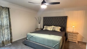 Un dormitorio con cama, ventilador de techo, mesita de noche, lámpara y un florero con ramas.