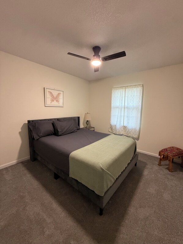 A bedroom with a bed, a ceiling fan, a window with curtains, a bedside lamp, and a small stool.