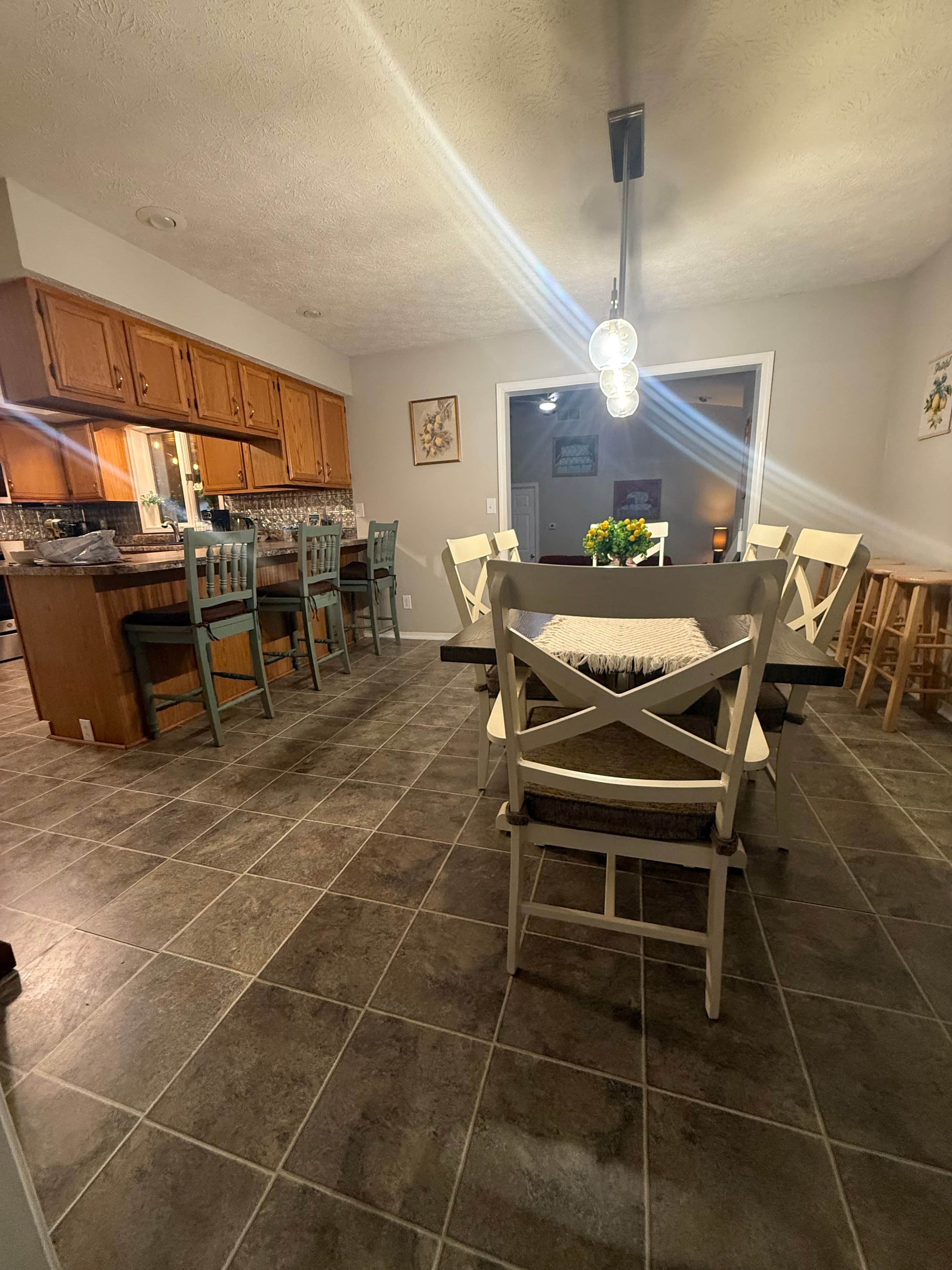 A dining area with a table and chairs, a kitchen in the background, and a ceiling light fixture.