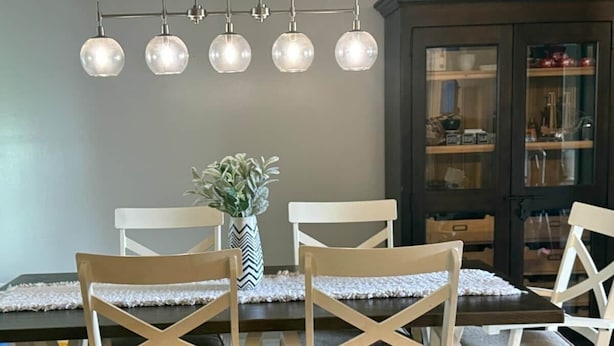 Un coin repas avec une table en bois foncé, des chaises blanches au dossier croisé, un vase avec des plantes vertes et un lustre moderne doté de cinq lampes sphériques.