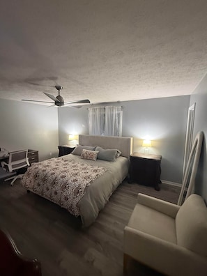 Een slaapkamer met een bed, twee nachtkastjes met lampen, een plafondventilator, een stoel en een bureau met stoel.