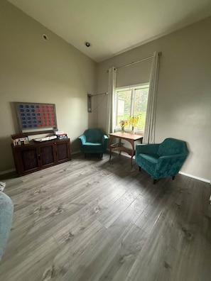 A room with a wooden floor, teal chairs, a small table with a vase, and a wall-mounted shelf with books.