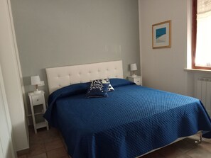 Un dormitorio con cama, mesita de noche, lámpara, almohada, un cuadro enmarco y ventana.