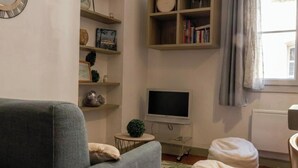 Una sala de estar con un sofá gris, un televisor, estanterías con libros y objetos decorativos, y una ventana con cortinas.