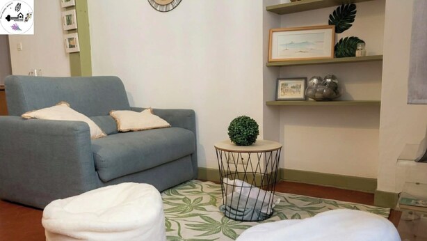 Una sala de estar con un sofá gris, dos otomanas blancas, una mesita redonda con una planta y una alfombra estampada.