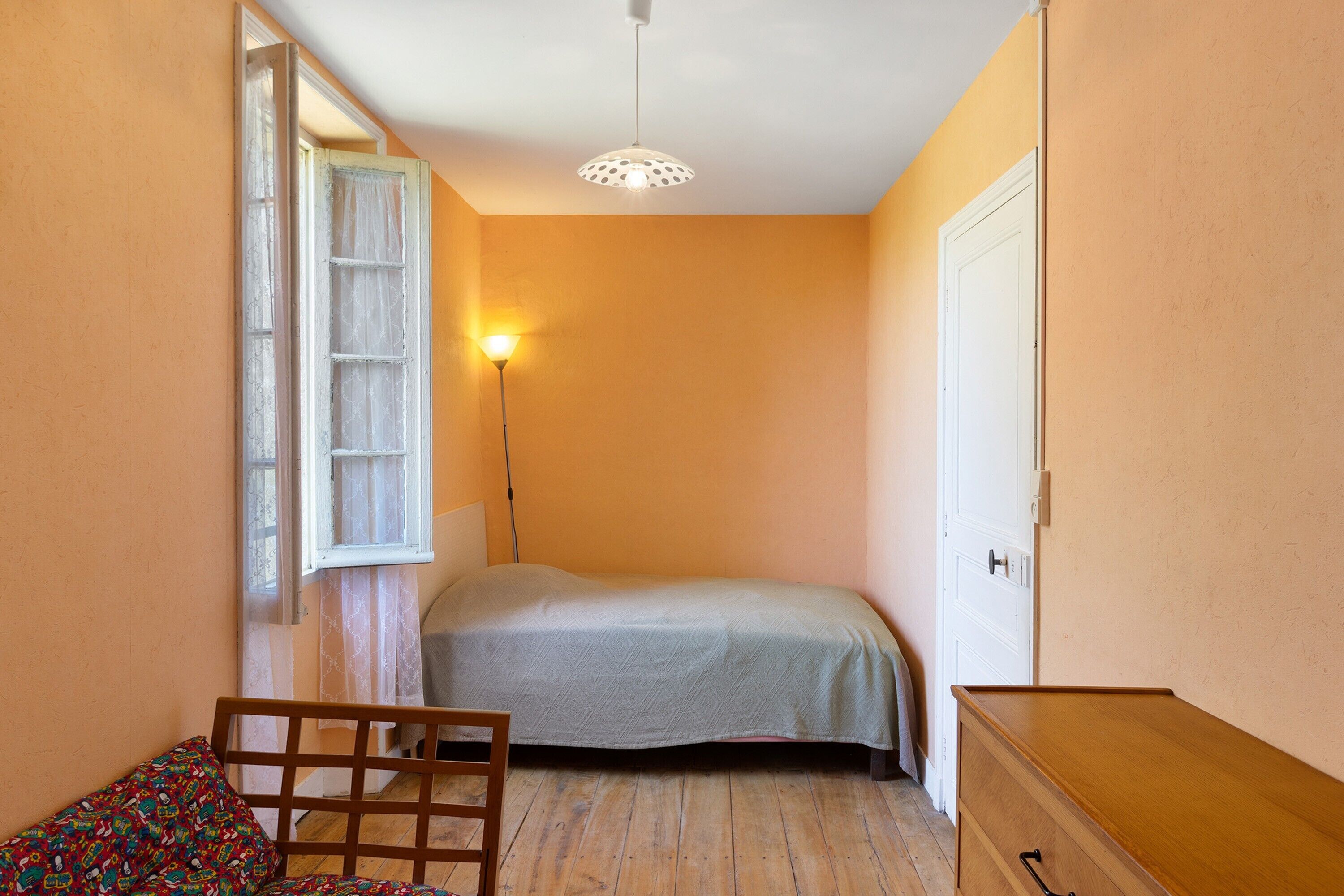 A bedroom with a bed, a chair, a dresser, a window with curtains, and a floor lamp.