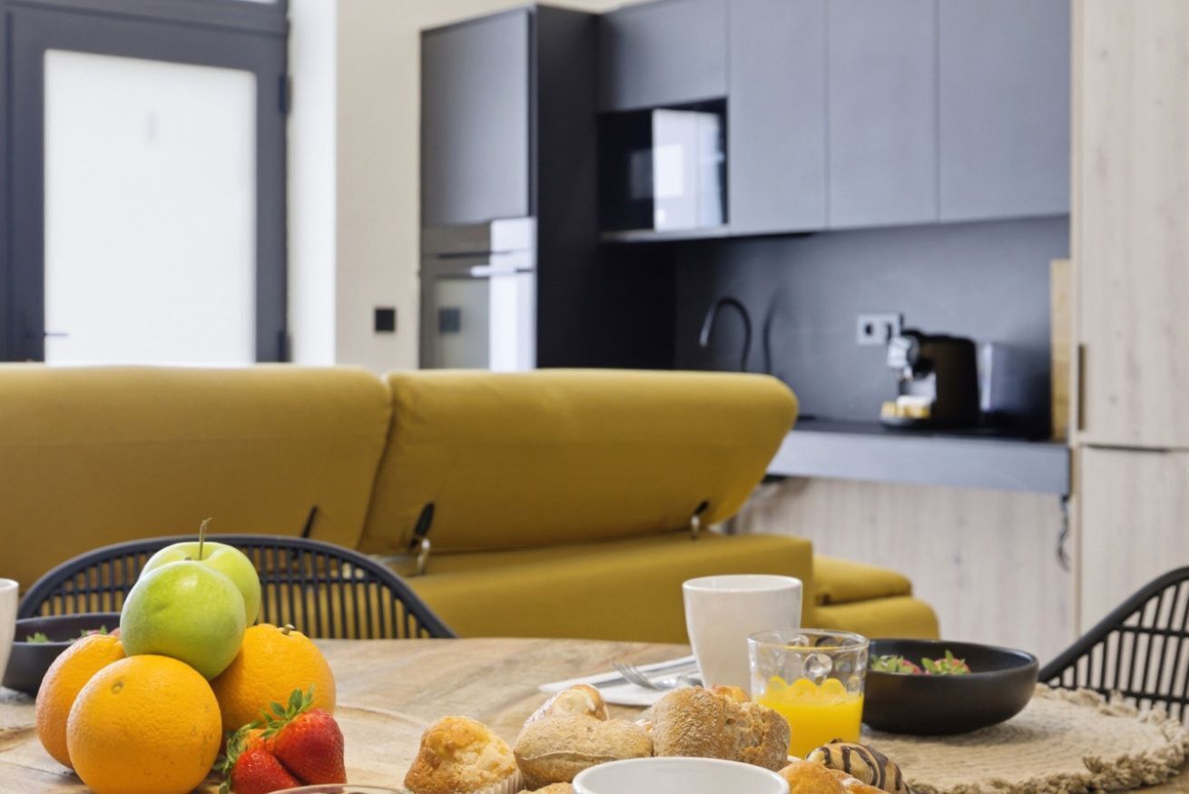 Ein Esstisch mit Früchten, Muffins und einem Glas Orangensaft in einer modernen Küche mit einer Couch und Schränken.
