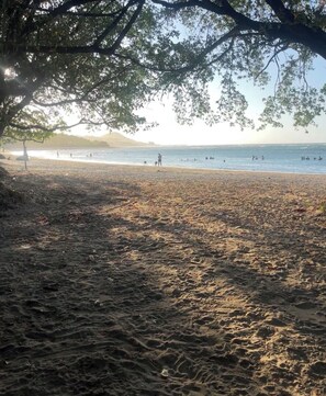 Ein Strand mit einem Baum, der Schatten spendet, feinem Sand und dem Meer im Hintergrund.