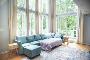 A room with a large window, a sectional sofa, a small wooden nightstand, and a patterned rug. - Five Bedroom Brookview Estate (Atlanta)