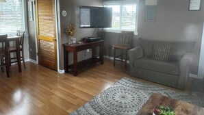 Una sala de estar con piso de madera, un sofá gris, un televisor, una mesa de comedor con sillas y un tapete estampado.